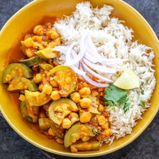 zucchini chickpea curry served with white rice in a yellow color bowl topped with sliced onion, cilantro and a lime wedge