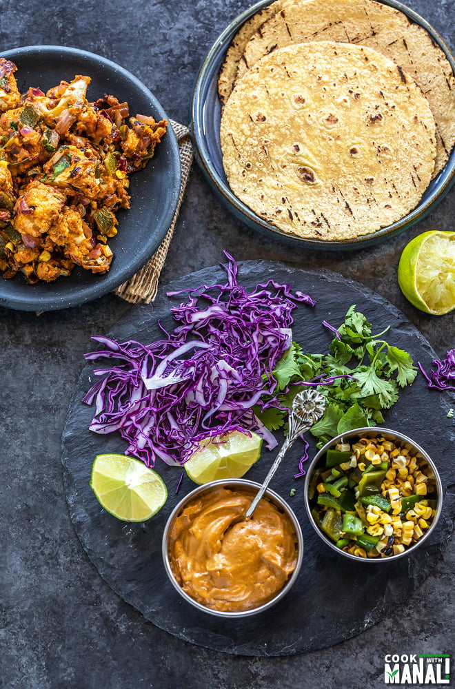 a plate with tortillas and another with cauliflower with 2 bowls filled with sauce and roasted corn and some purple cabbage, cilantro and lime placed on the sides