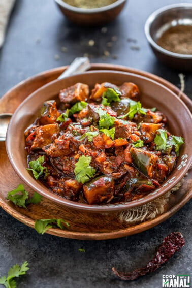 eggplant curry served in a brown bowl, garnished with cilantro and bowls of spices placed in the background