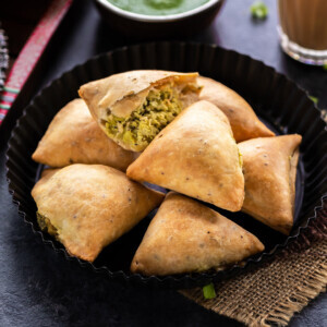 7 baked samosa arranged on a plate with glass of chai on the side and bowl of cilantro chutney in the background