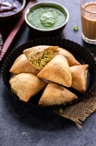 7 baked samosa arranged on a plate with glass of chai on the side and bowl of cilantro chutney in the background