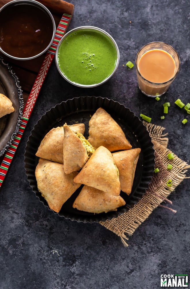 plate of baked samosa and bowls of chutney in the background and a glass of chai on the side