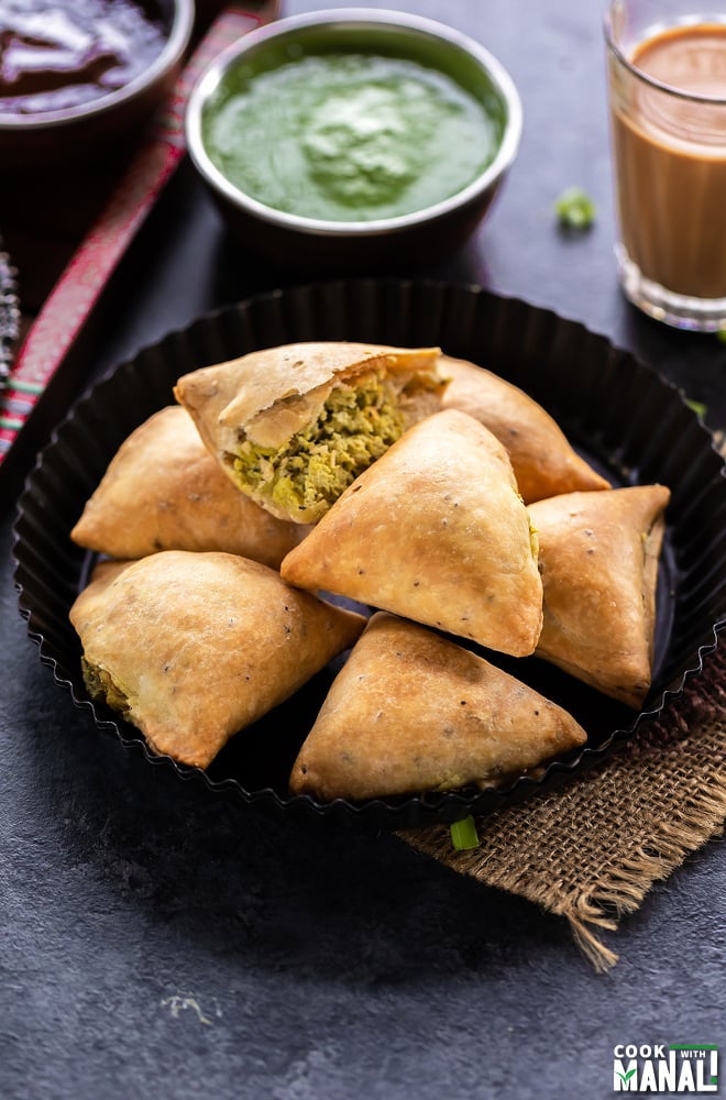 7 baked samosa arranged on a plate with glass of chai on the side and bowl of cilantro chutney in the background 