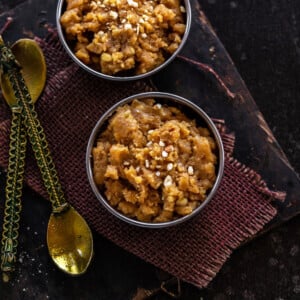 besan halwa served in a copper bowl with two golden spoons placed on the side and another bowl of halwa in the back