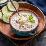 bowl of cucumber raita in a blue bowl garnished with mint leaves and spices and few round slices of cucumber in the background