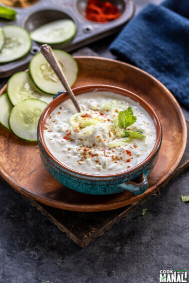 bowl of cucumber raita in a blue bowl garnished with mint leaves and spices and few round slices of cucumber in the background
