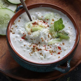 bowl of yogurt with grated cucumber and mint leaves and some spices sprinkled on top