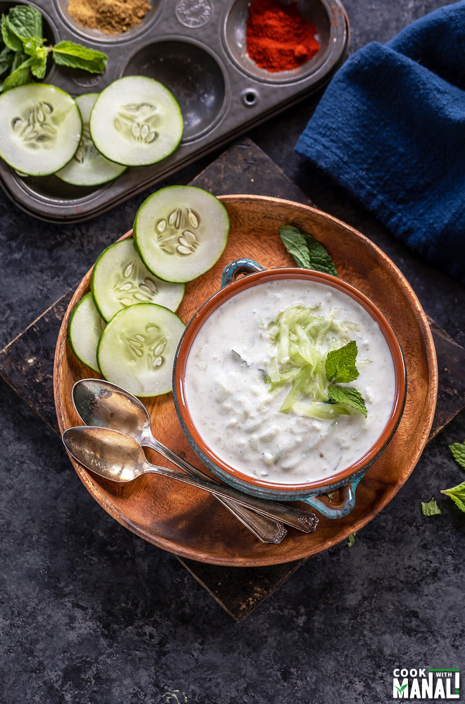 overhead shot of cucumber raita in a bowl garnished with mint and slices of cucumber and some spices placed in the background
