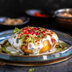 raj kachori placed on a plate topped with yogurt, chutney, pomegranate, cilantro and bowl of spices placed in the background