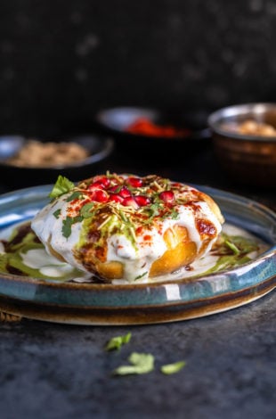 raj kachori placed on a plate topped with yogurt, chutney, pomegranate, cilantro and bowl of spices placed in the background