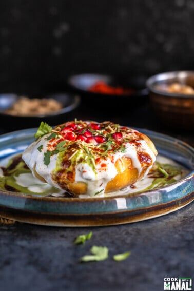 raj kachori placed on a plate topped with yogurt, chutney, pomegranate, cilantro and bowl of spices placed in the background