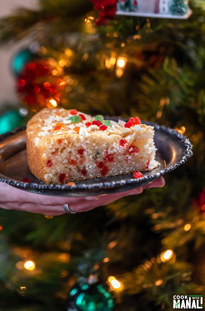 a hand holding a plate of cake in front of a Christmas tree
