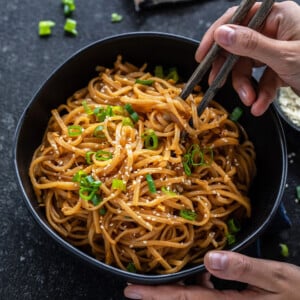 pair of hands holding a bowl of noodles with one hand holding a pair of chopsticks