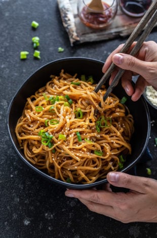 pair of hands holding a bowl of noodles with one hand holding a pair of chopsticks