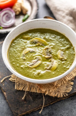 mushroom and spinach curry served in a white bowl with onions, tomatoes placed in the background