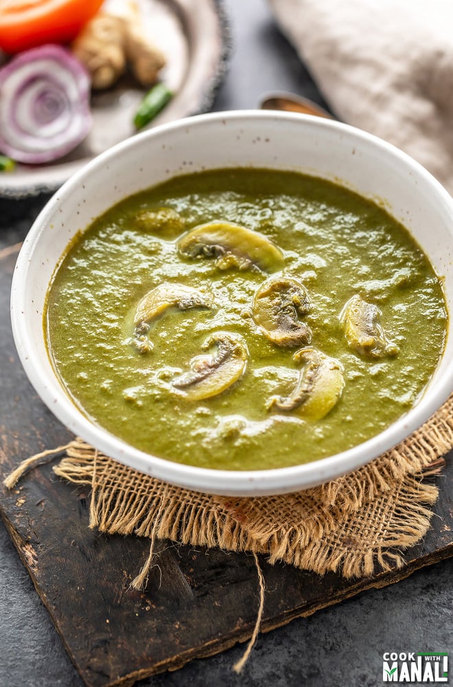 mushroom and spinach curry served in a white bowl with onions, tomatoes placed in the background