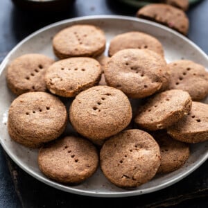 plate full of ragi cookies with bowl of jaggery and a glass of chai in the background