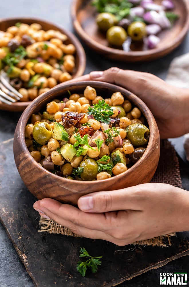 pair of hand holding a bowl of chickpea salad served in a wooden bowl