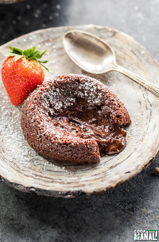 chocolate lava cake placed on a plate with molten chocolate flowing through the center and a strawberry placed in the back