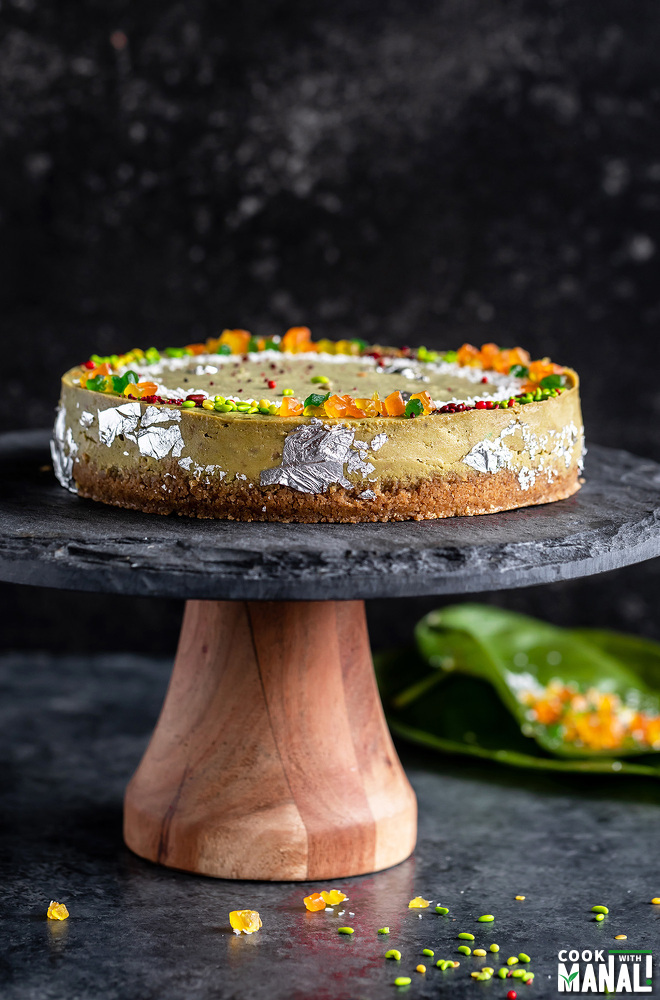 a green color cheesecake on a black stand topped with tutti frutti, edible silver leaves