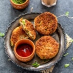 3 veg cutlet placed on a plate with a bowl of ketchup and bowl of chutney and glass of chai in the background