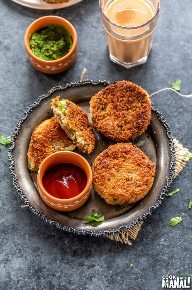 3 veg cutlet placed on a plate with a bowl of ketchup and bowl of chutney and glass of chai in the background