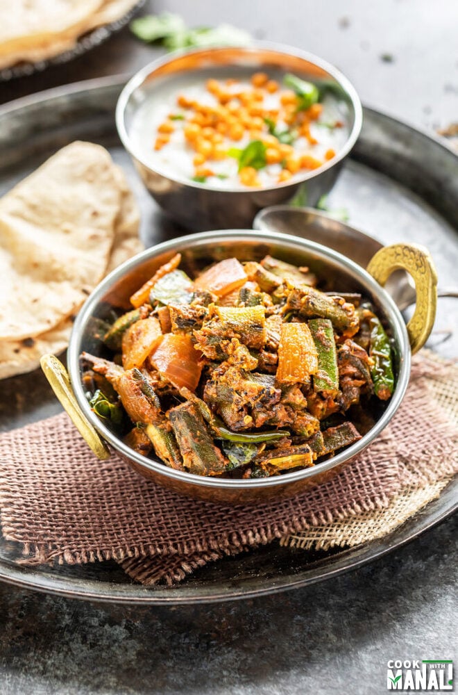 bhindi served in a copper kadai with rotis and a bowl of boondi raita placed in the background