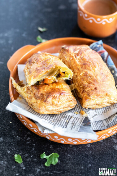 a veg puff cut into half to reveal the filling and a cup of chai placed in the background