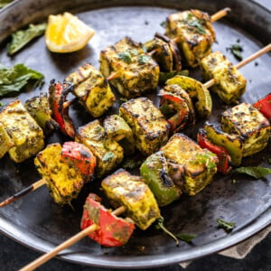 3 skewers of hariyali paneer tikka placed on a round plate with a lemon wedge on the side