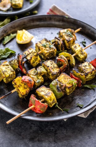 3 skewers of hariyali paneer tikka placed on a round plate with a lemon wedge on the side