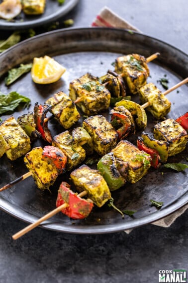 3 skewers of hariyali paneer tikka placed on a round plate with a lemon wedge on the side