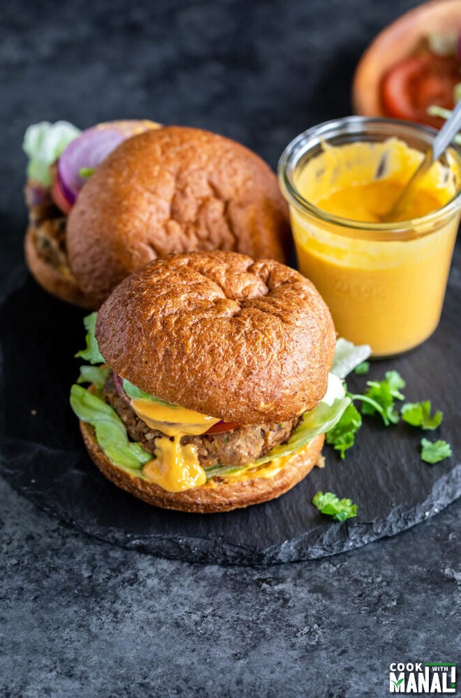 2 burgers placed on a black board with a yellow sauce jar in the back