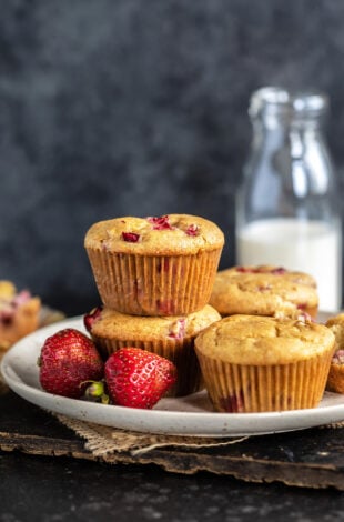 2 strawberry muffins stacked together with 2 more placed on the side and fresh strawberries placed in the front of the plate