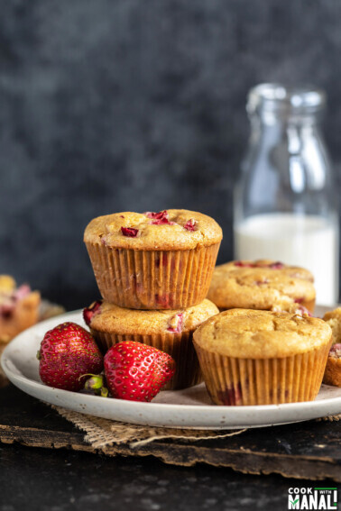 2 strawberry muffins stacked together with 2 more placed on the side and fresh strawberries placed in the front of the plate