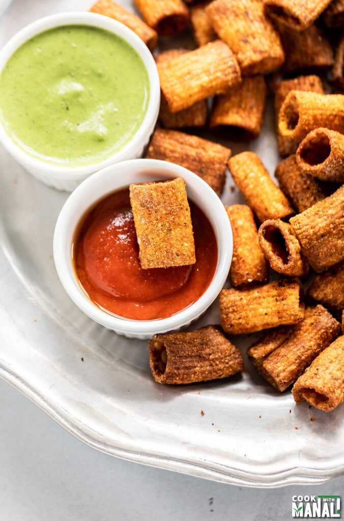 pasta chips served in a plate with 1 bowl of chutney and another bowl of hot sauce