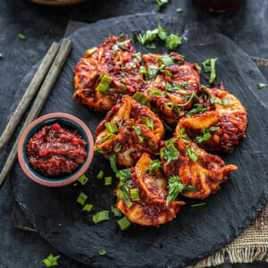 momos topped with cilantro, green onions arranged on a black board with a bowl of hot sauce and pair of chopsticks placed on the side