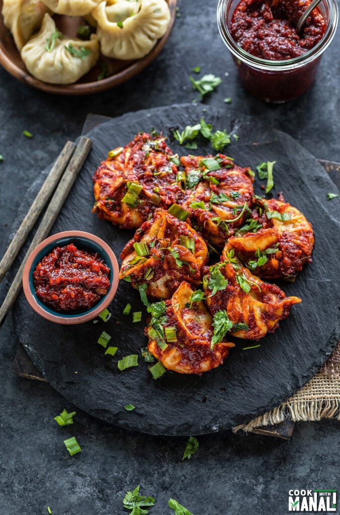 momos topped with cilantro, green onions arranged on a black board with a bowl of hot sauce and pair of chopsticks placed on the side