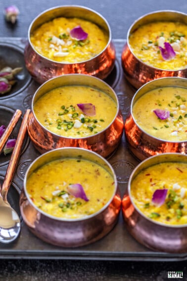 6 copper bowls arranged on a tray, filled with rabdi and topped with nuts and rose petals