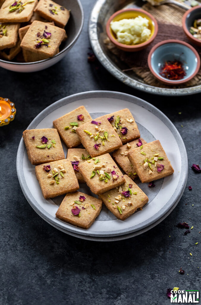 cookies topped with rose petals and pistachios placed on a plate with small bowls containing saffron placed in the background