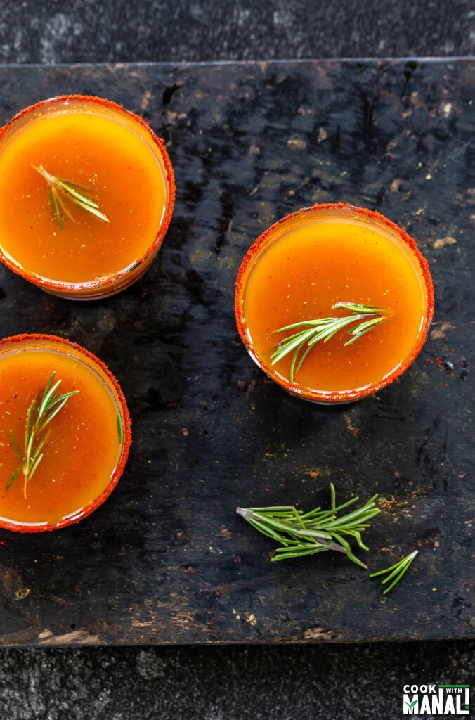 3 small glasses with orange color drink and garnished with rosemary