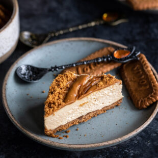 slice of biscoff cheesecake placed on a blue color plate with 2 biscoff cookies and a spoon placed on the side