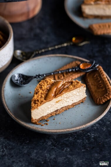slice of biscoff cheesecake placed on a blue color plate with 2 biscoff cookies and a spoon placed on the side