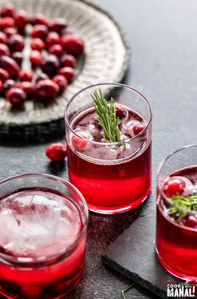 three glasses filled with red color drinks and plate of cranberries in the back