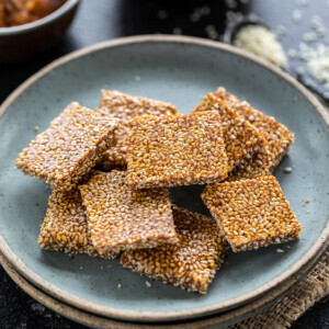 til chikki squares placed in a blue plate with bowls of jaggery and sesame seeds placed in the background