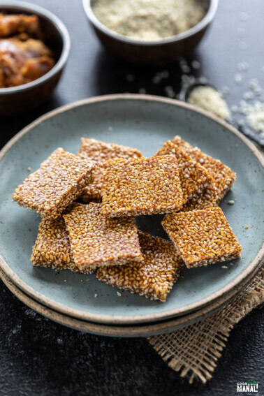 til chikki squares placed in a blue plate with bowls of jaggery and sesame seeds placed in the background