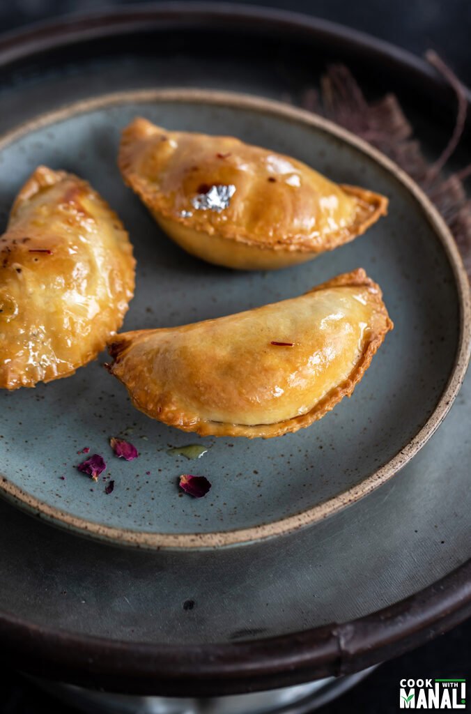 3 pieces of gujiya placed on a blue plate with some crushed rose petals on the side