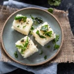 3 pieces of rava dhokla served on a blue plate with bowl of chutney in the background