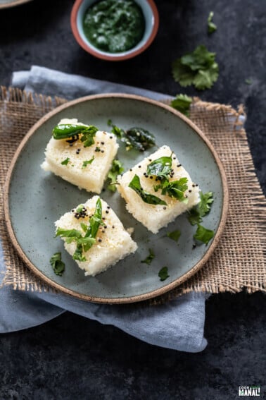 3 pieces of rava dhokla served on a blue plate with bowl of chutney in the background