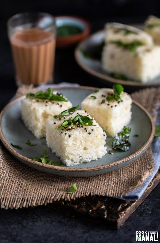 3 pieces of sooji dhokla served on a plate with glass of chai and more dhokla in the background