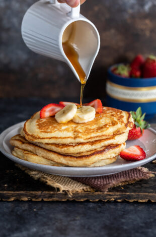 maple syrup being poured over a stack of pancakes topped with strawberries and banana